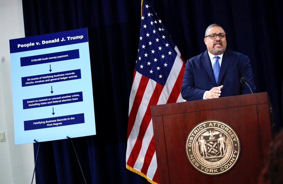 A Black man wears a blue suit and stands at a New York County lectern next to a poster that says 'People v. Donald J. Trump' and in front of an American flag.