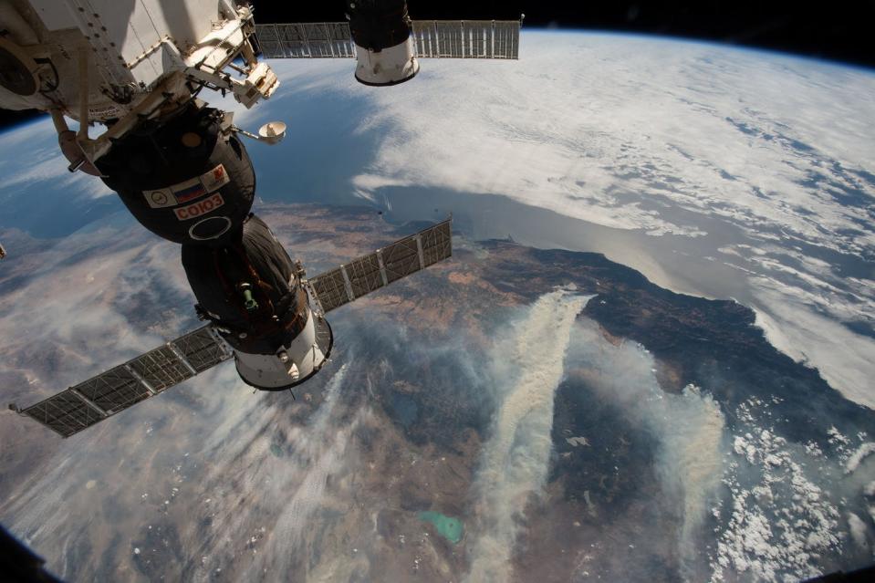 An image shot from the space station showing part of the space station and land below with smoke from wildfires.