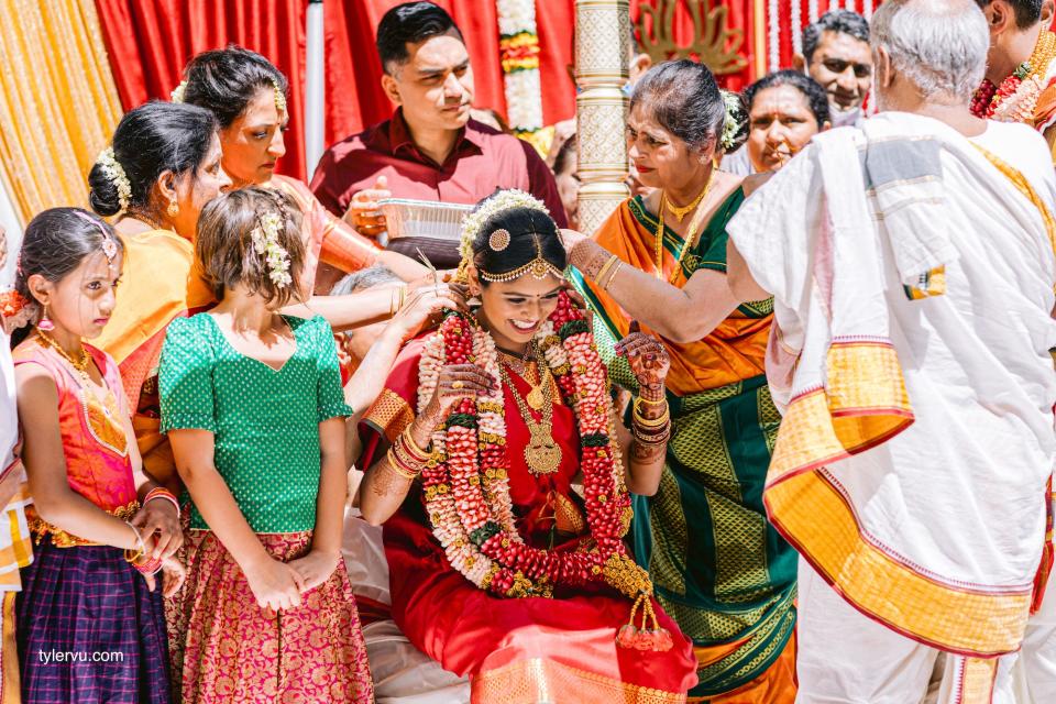 A bride in a red lehnga smiles as her family places a floral necklace around her.