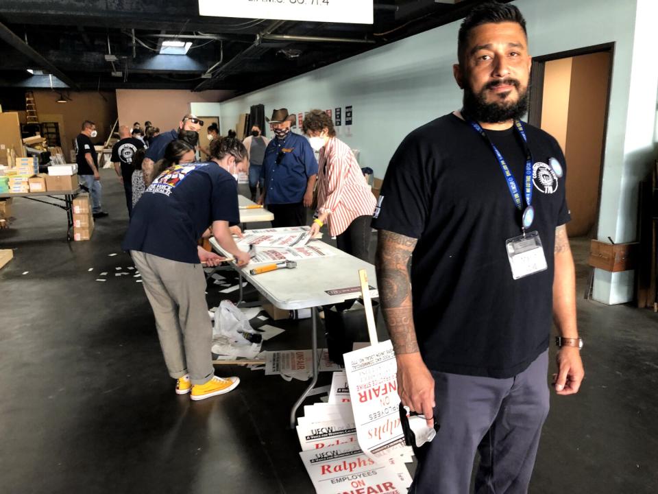 A man posing for a photo. Behind him are people working at a white table, cutting out signs and organizing boxes.