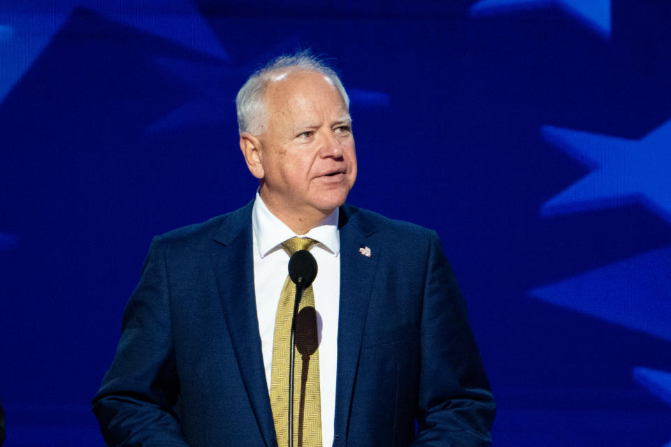 Tim Walz standing at a podium, wearing a suit and tie, with a blue starry backdrop behind him