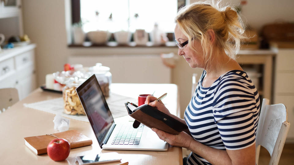 Beautiful senior woman with laptop at home at the kitchen table, writing something into her calendar.