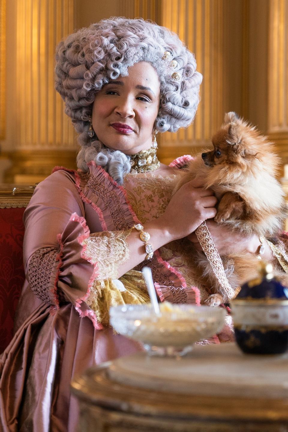 Woman in period costume with wig holds a small dog, seated in an ornate room