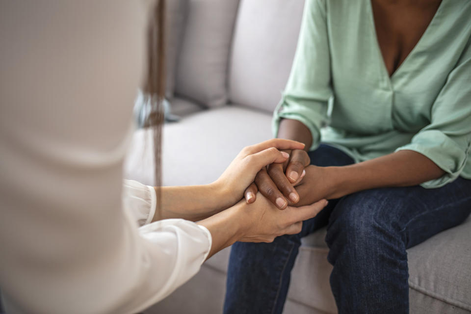 A person in a green blouse and jeans sits on a couch holding hands with another person in a white shirt, providing comfort and support