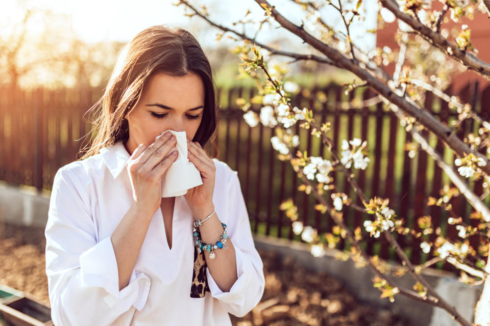 A young woman standing near a blooming tree holds a tissue to her nose.