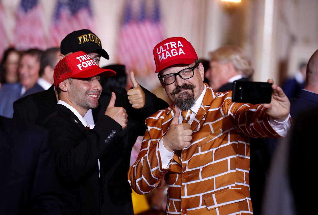 Three Trump supporters group to take a selfie at Mar-a-Lago, two wearing red baseball caps marked Trump Forever and Ultra Maga, and one wearing an orange jacket printed like a brick wall.