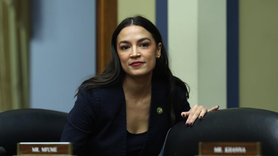 alexandria ocasio cortez preparing to sit down behind a panel with multiple chairs