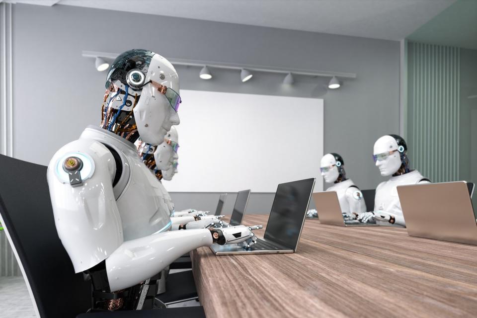 Several humanoid robots type on laptops while sitting at a long table in a conference room.