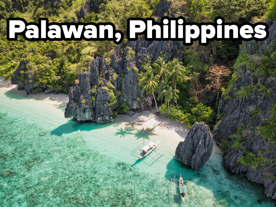 Tall rock formations jutting out of the ground, with clear ocean waters, and a white sand beach. Tall palm trees towering over the sands, and a few traditional boats floating near the shore.