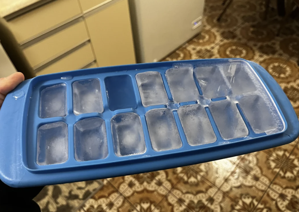 An ice cube tray with many compartments containing half-melted ice cubes is held in one hand over a patterned kitchen floor