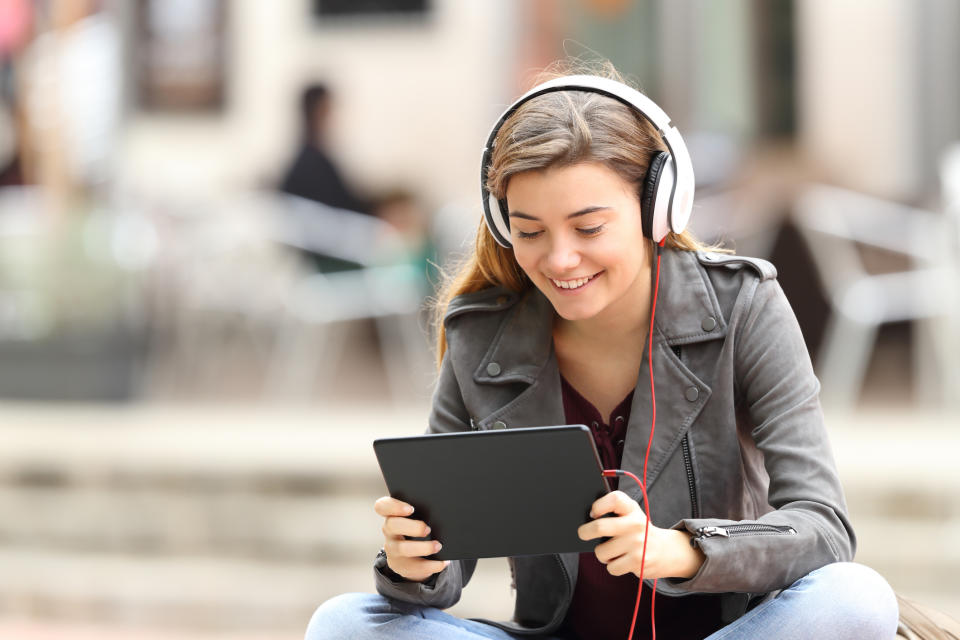 Girl sitting wearing headphones smiling and looking at a tablet.