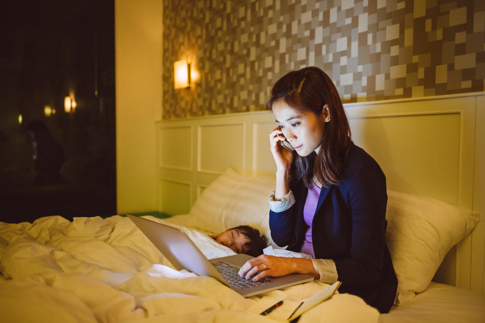 Pretty young mom talking on the phone while working with the laptop on the bed with little girl sleeping soundly beside her