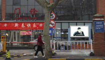 An elderly woman with a face mask walks past the entrance to the outpatient ward with a video display showing prevention methods in Shanghai on Monday, Jan. 27, 2020. The government of Shanghai, a metropolis of 25 million people and a global business center, extended the holiday by an additional week within the city to Feb. 9. It ordered sports stadiums and religious events closed. (AP Photo/Erika Kinetz)