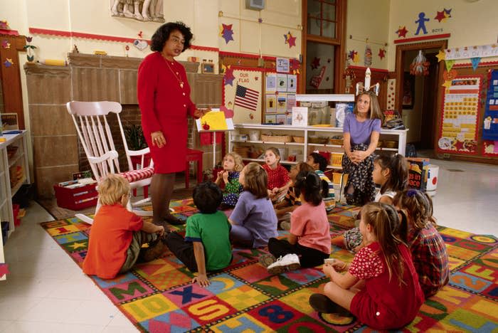 A teacher standing in a classroom, with children sitting on the floor looking at her. She's holding a book