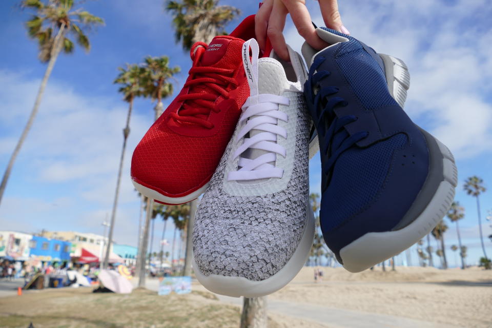 A person holds three Skechers shoes at the beach.