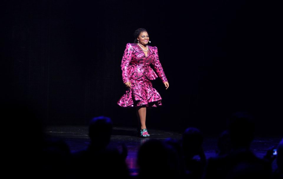 da'vine joy randolph walks across a stage while lit in a spotlight, she wears a pink cheetah patterned skirt suit with ruffles, a beaded necklace and a microphone, she smiles as people sit in the foreground facing her