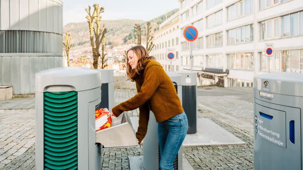 A woman places a small red and white bag of trash inside of a squat gray metal box built into the cobblestone streets of Bergen, Norway.