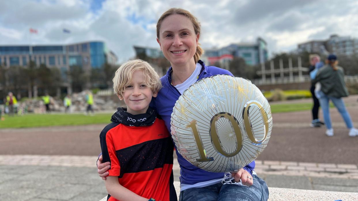Verity hugs Xander with one arm as they sit on a bench looking at the camera while Verity also holds a balloon with 100 on it