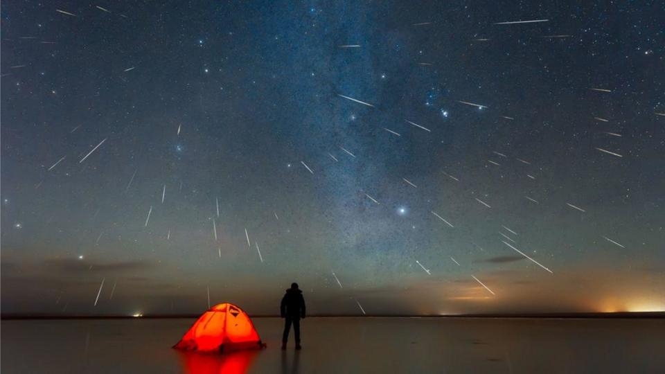 La silueta de un hombre al lado de una carpa roja observando una lluvia de meteoros 