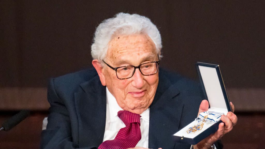 henry kissinger smiles and holds open a box with a medal inside, he wears a navy suit jacket, whit shirt and red tie