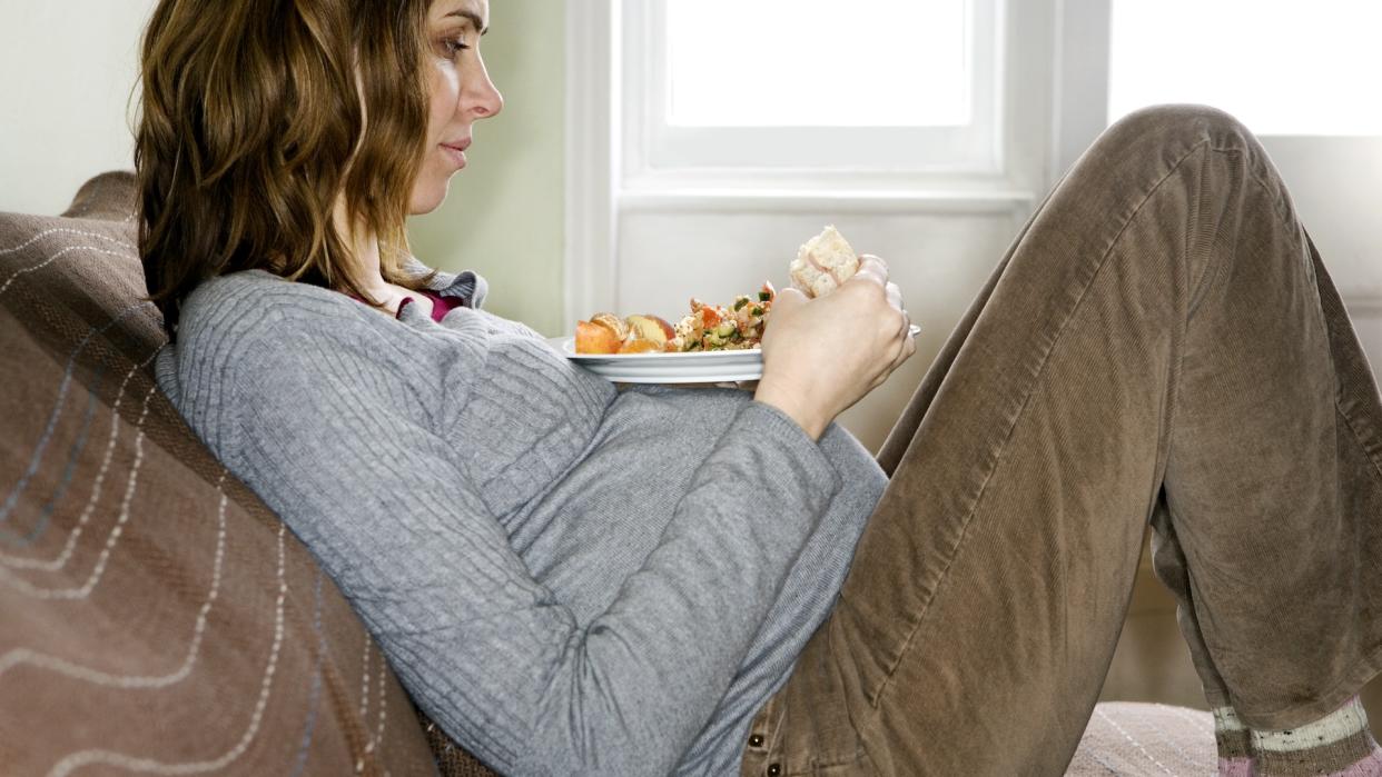  A pregnant woman sitting on a couch with her feet propped up and a plate of food resting on her belly. 