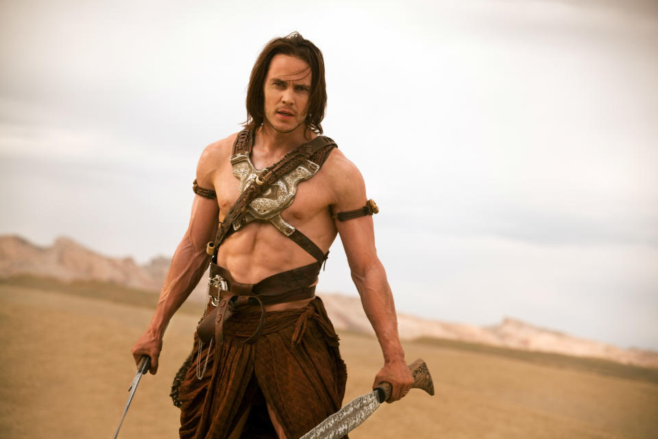 A person with long hair and muscular physique is wearing warrior-like attire, holding a sword, and standing in a desert setting