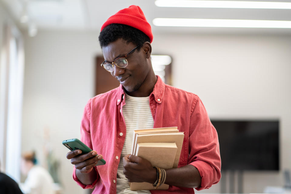 Smiling African American student studying on smartphone and reading sms message while studying in library, selective focus.  Black male teacher holding stack of books using planning app on mobile device