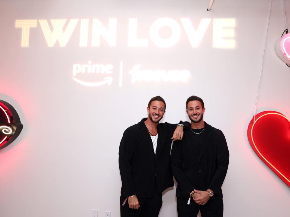 samer and samir akel in black clothes, smiling under a sign that reads twin love