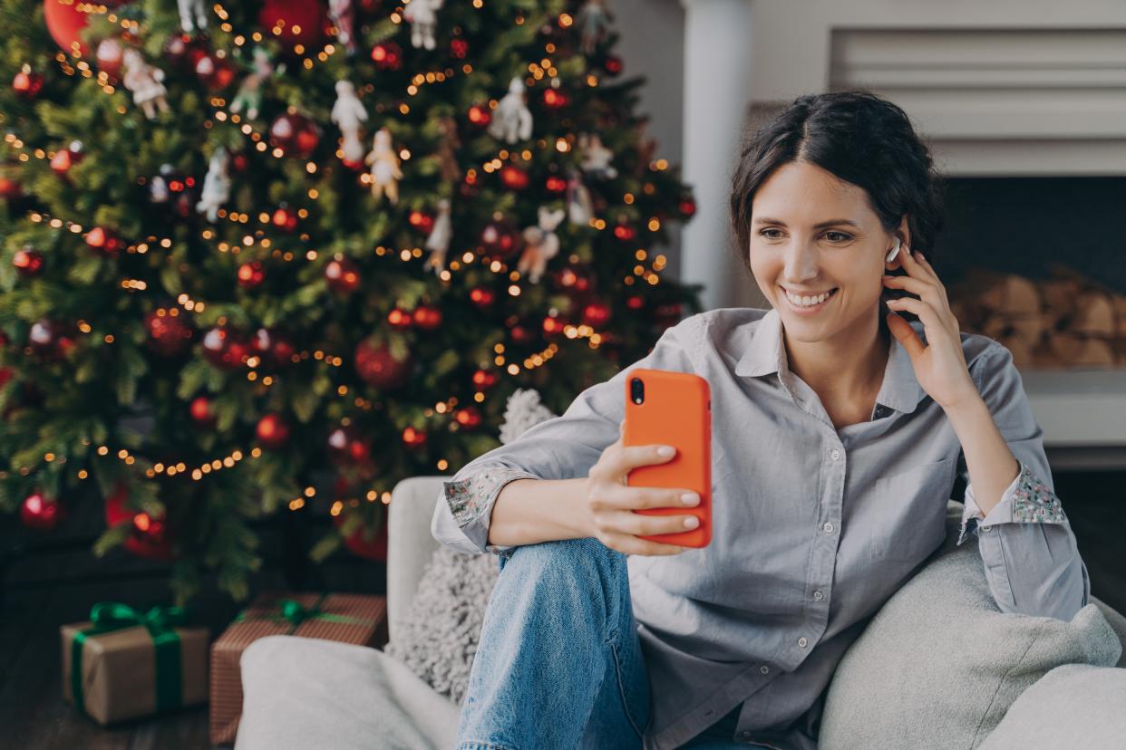 Smiling happy hispanic woman in wireless earphones holding phone, sitting near Christmas tree at home, looking at smartphone screen and making video call, using mobile device during winter holidays