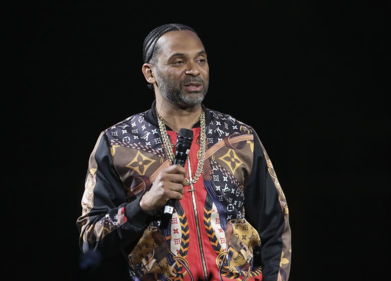A man in a patterned jacket and gold necklace speaking into a microphone
