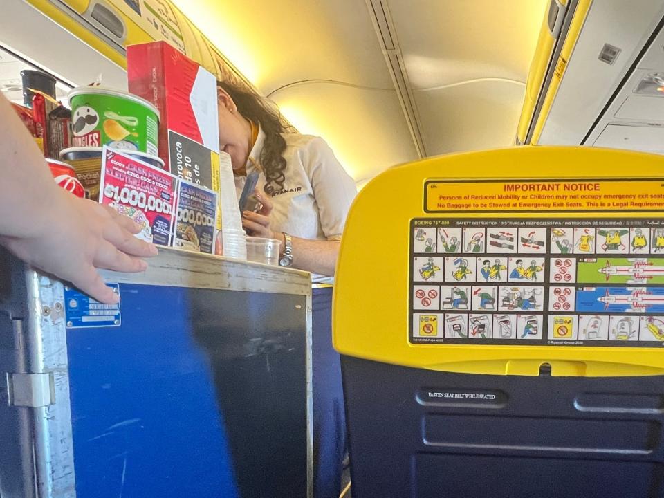 Two Ryanair flight attendants push a trolley down the aisle, with pringles and scratch cards and cigarettes on top, from the aisle seat's point of view.