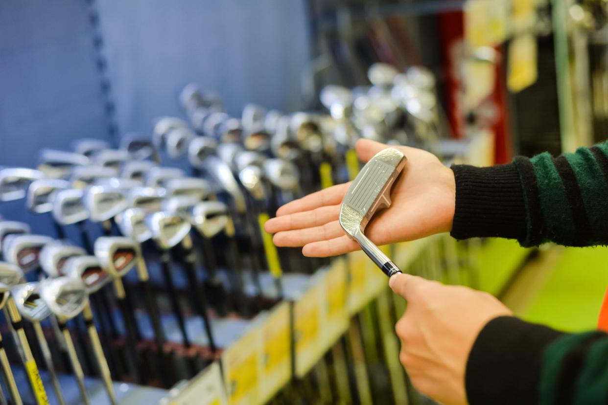 Focus on two male hands holding a left-handing golf club, with the clubhead on the palm of the right hand, with a blurred background of several rows of golf clubs standing up for sale in sporting store