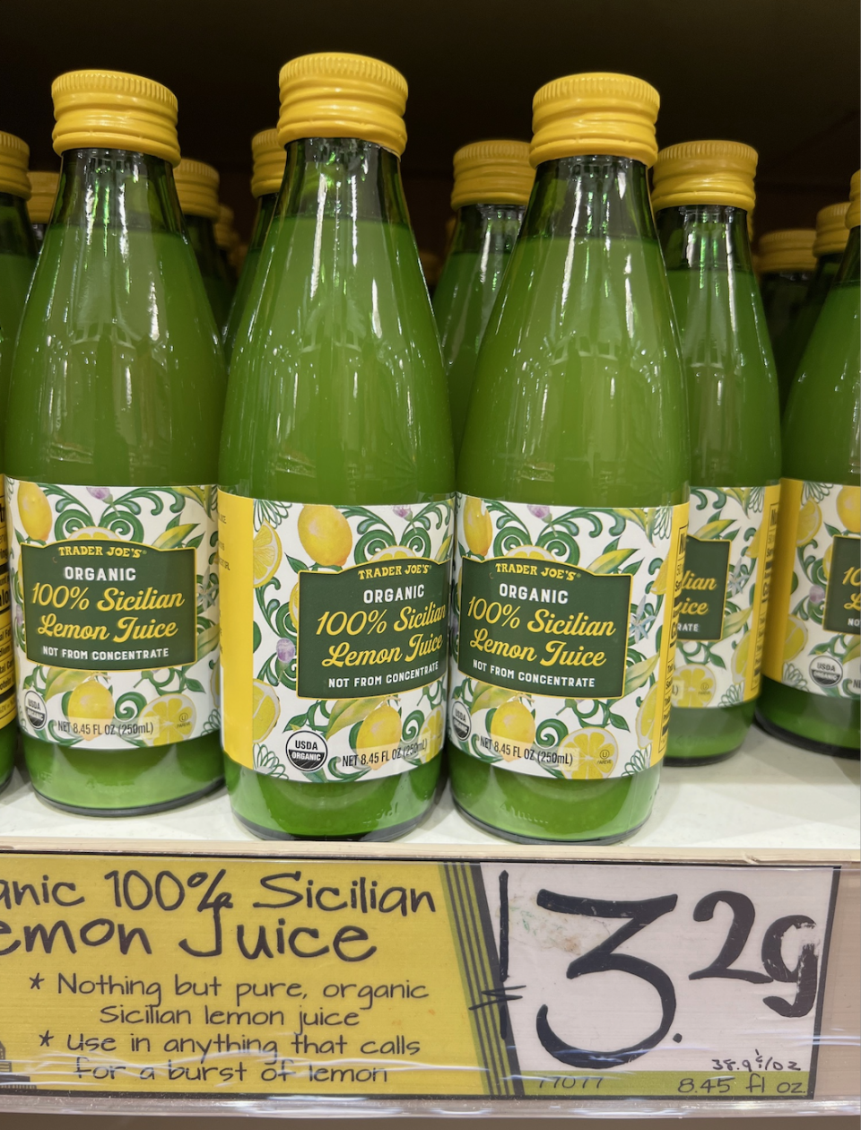 Several green bottles of Trader Joe's Organic 100% Sicilian Lemon Juice are displayed on a store shelf with a sign showing the price of $3.29