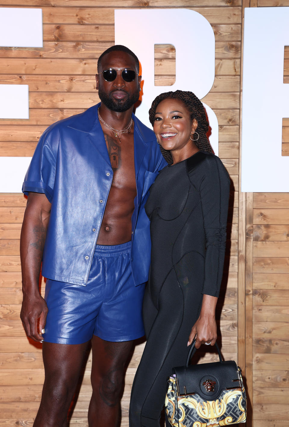 Gabrielle smiles as she takes a picture with her husband Dwyane at an event. She is wearing a body suit and he's wearing leather shorts and a matching open shirt