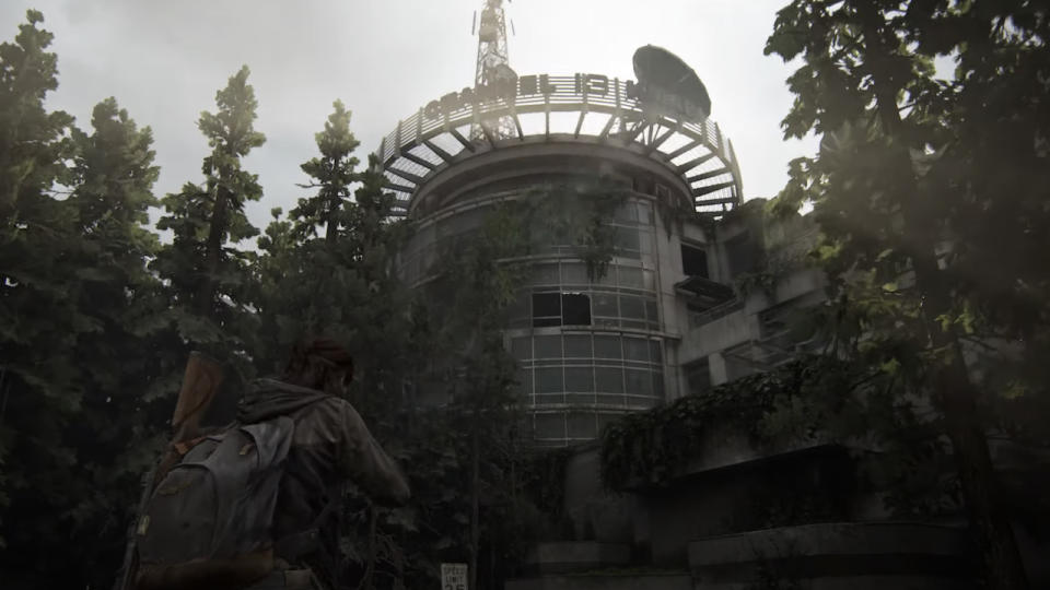 Ellie standing outdoors and looking up through the trees at the Channel 13 building in The Last of Us 2