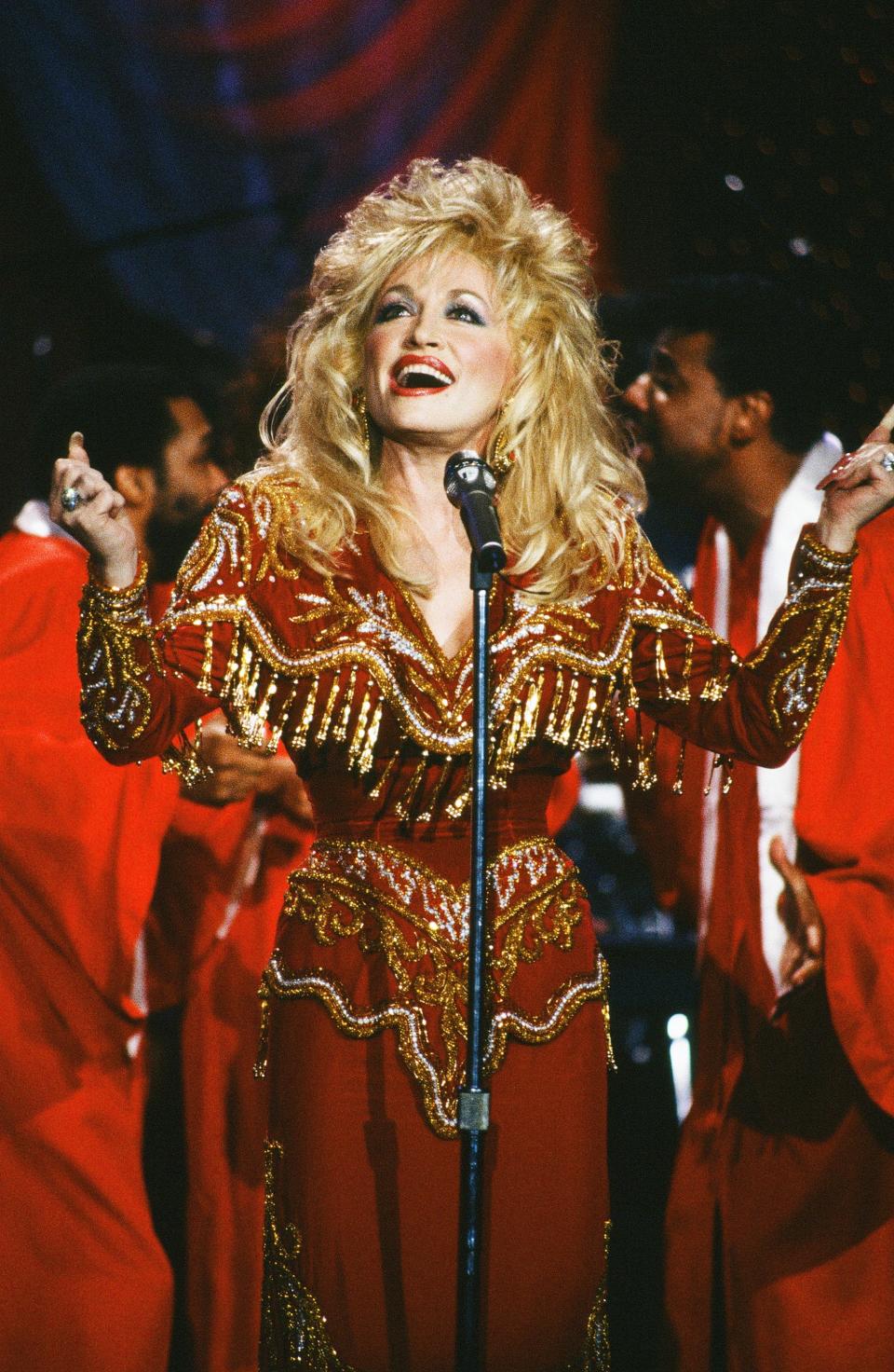 Dolly Parton singing in a red long sleeve gown covered in gold beads and tassels.