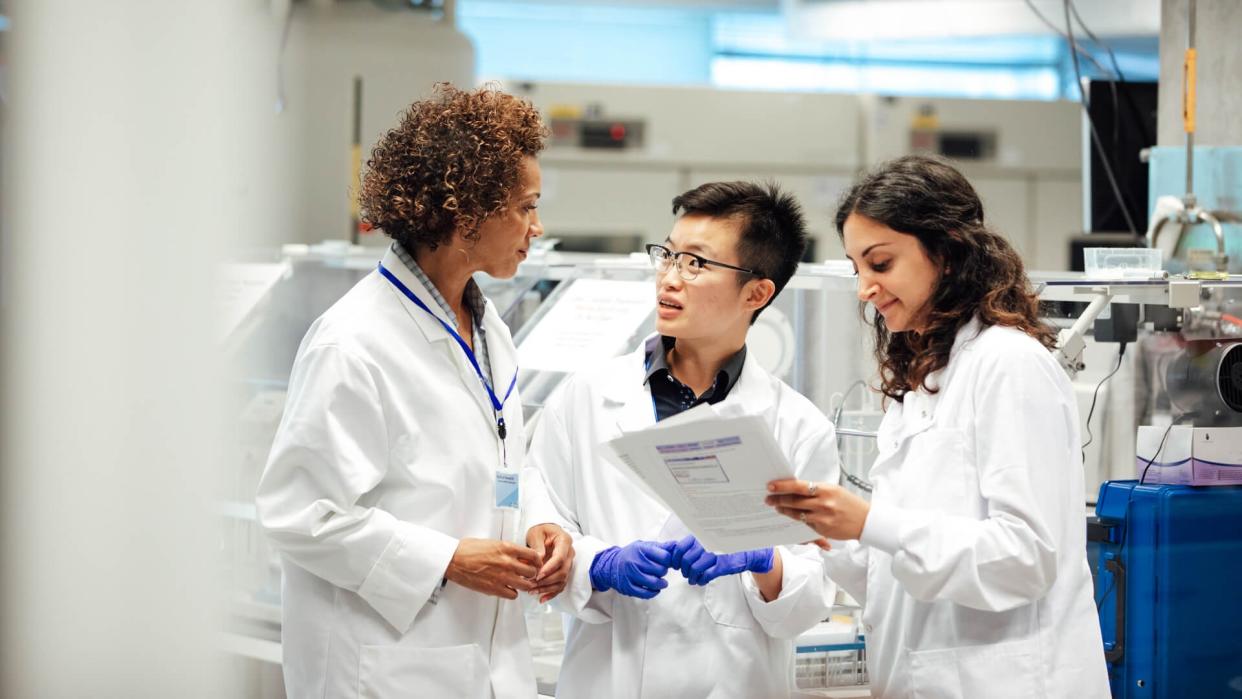 Diverse mix of technicians, wearing lab coats, debate working procedure, in a disease research facility.