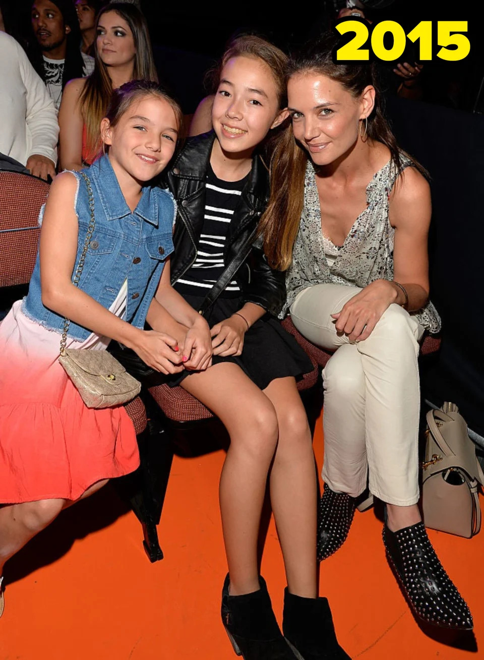 Katie Holmes sits with Suri Cruise and a friend. Katie is in white pants and studded boots, Suri wears a denim vest and ombre dress, and the friend wears a leather jacket