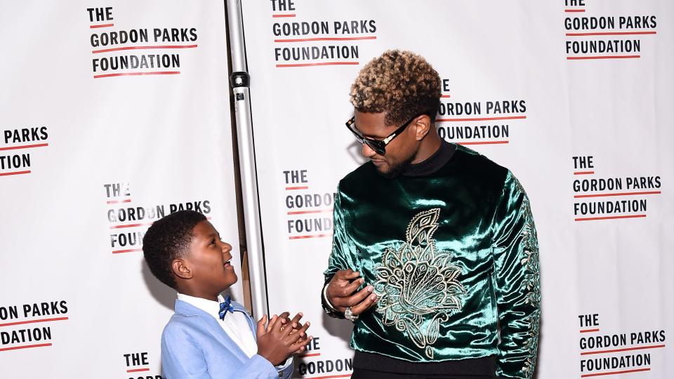usher, wearing a blue and black designed shirt, and his son naviyd ely, wearing a blue tuxedo, speak in front of a promotional wall for the gordon parks foundation