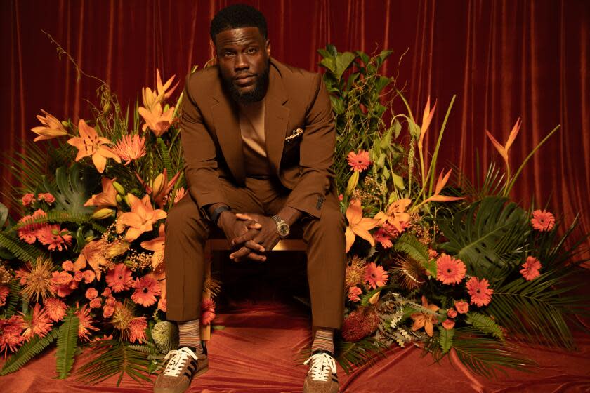 Kevin Hart sits on a chair surrounded by orange flowers and leans forward onto his knees