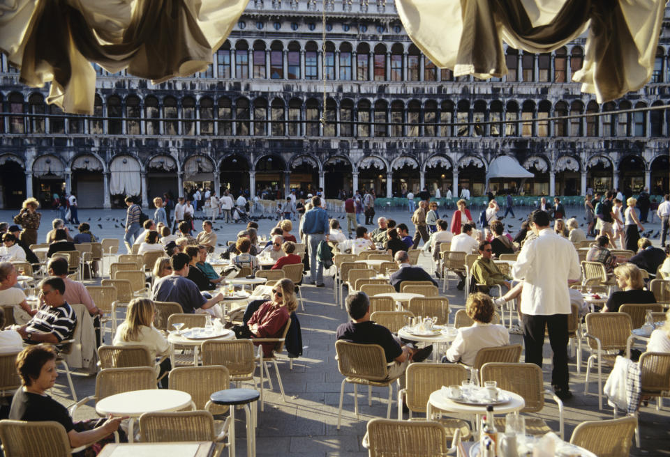 People sitting at an outdoor café in a busy square with an ornate building in the background