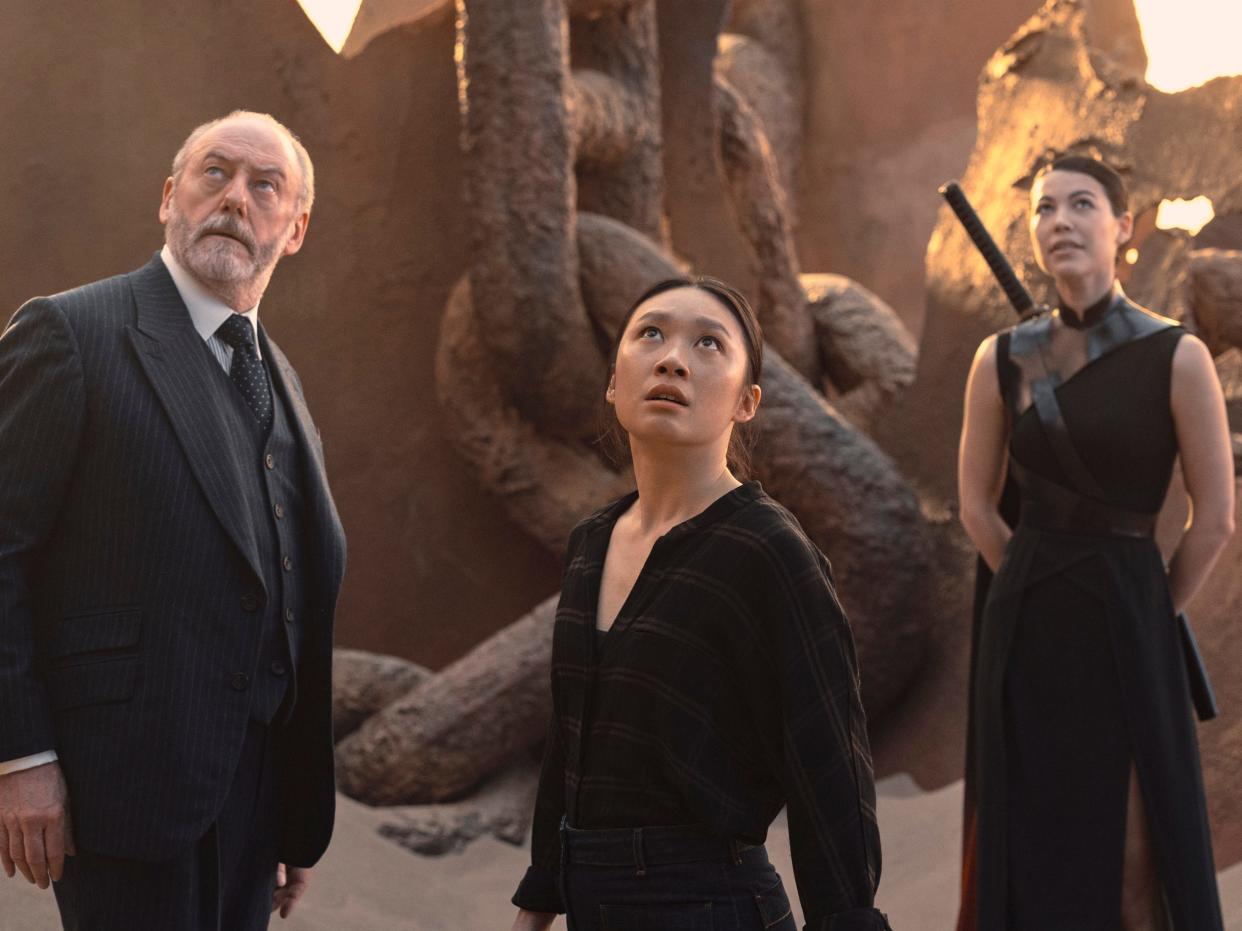 wade, jin cheng, and sophon in 3 body problem, standing in a sandy landscape and looking up at something in the sky. wade is a middle aged man in a suit, jin cheng is a young woman in casual clothing, and sophon is a young woman with a katana strapped to her back and in a flowing dress