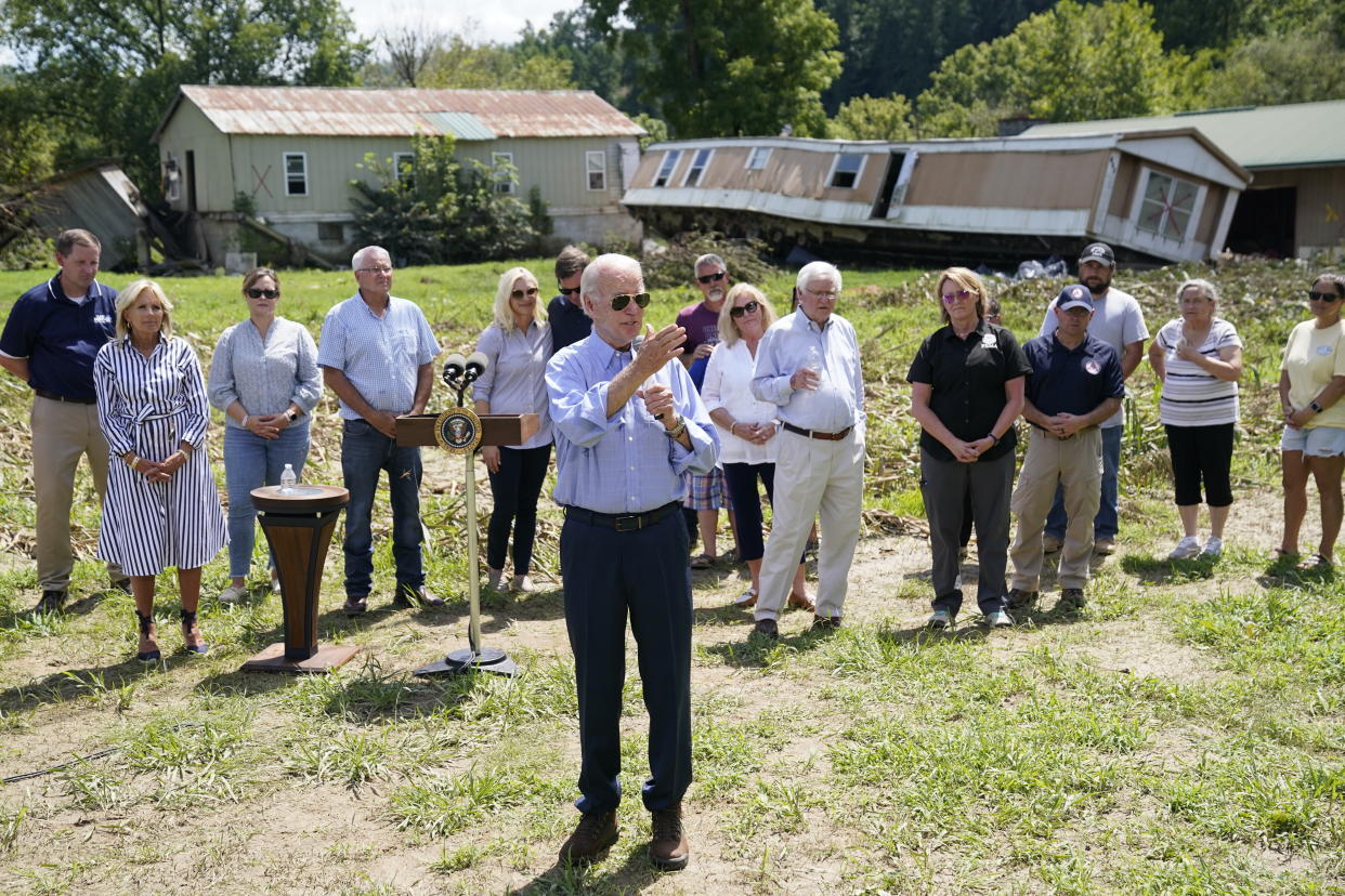 President Biden speaks while people stand around him and a flood-damaged home is seen in the background.