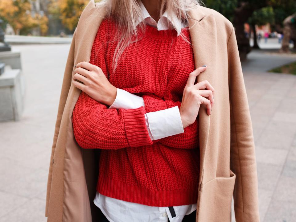 Woman wearing tan coat, red sweater, and white button-up blouse.