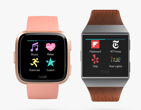 The Fitbit Versa, with rounded edges and in pink, displayed on the left. On the right is the older Ionic, with square edges, silver trim, and brown leather band.