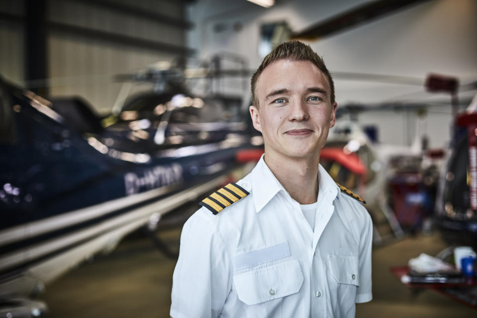 Pilot in uniform smiling in aircraft hangar, helicopter in background