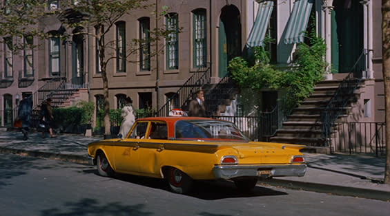 A yellow taxi is parked on a quiet city street lined with brownstones and greenery, with people walking on the sidewalk in the background