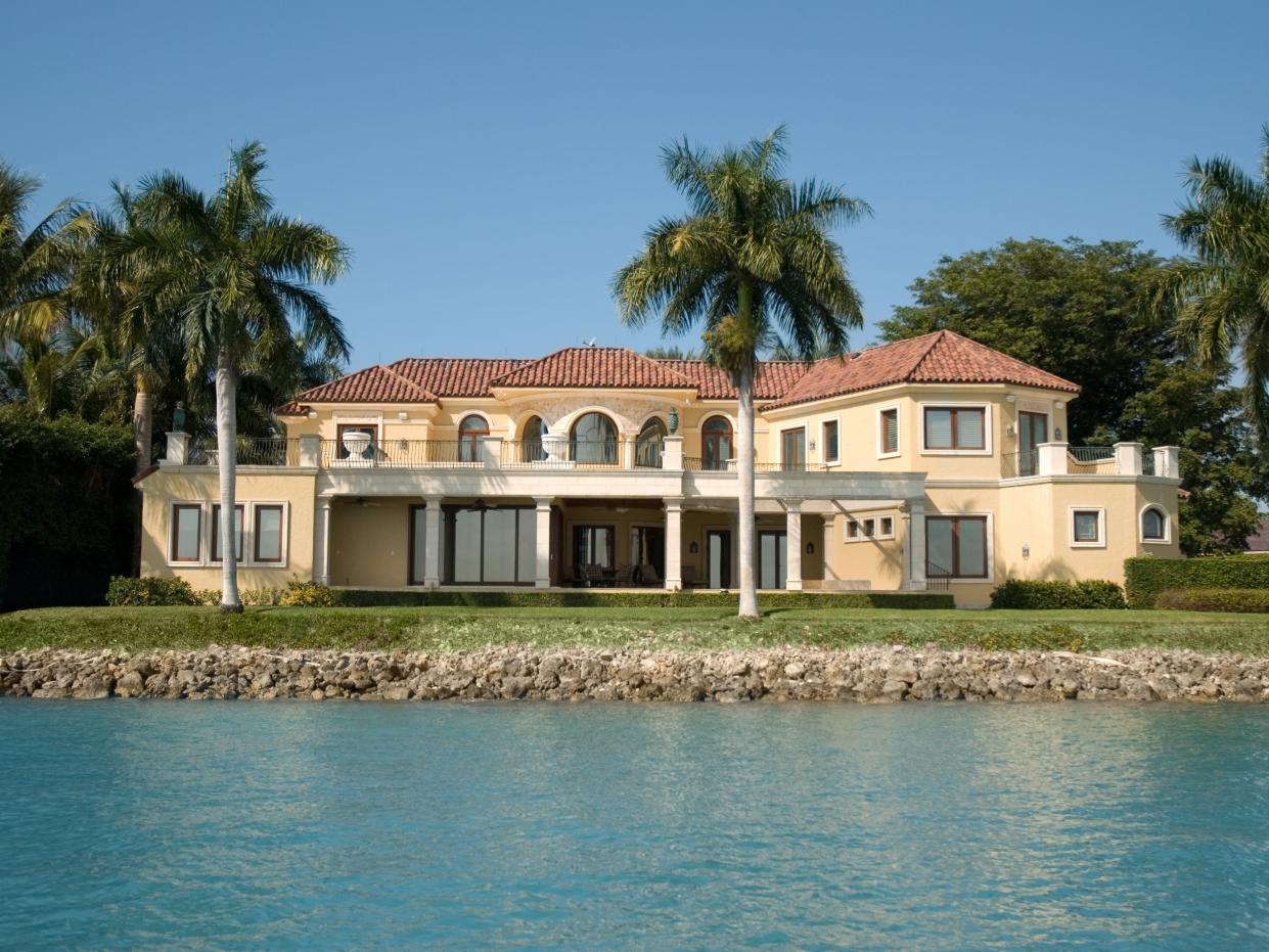 Empty sprawling mansion on manicured lawn with palm trees in the background.