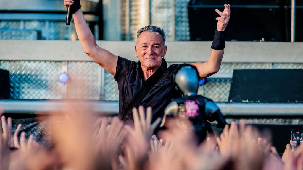 bruce springsteen raising both arms in the air and holding a microphone in front of a concert crowd
