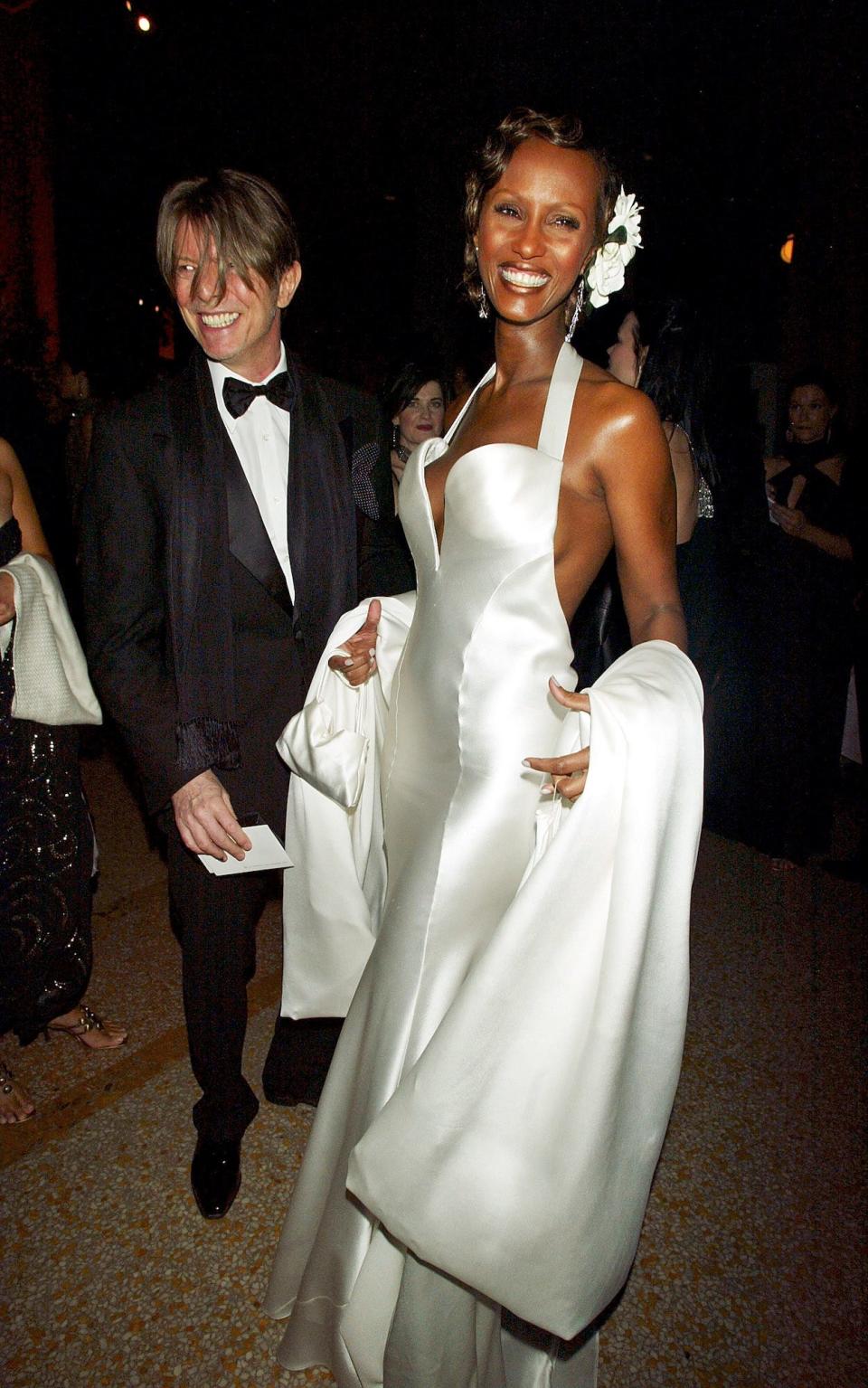 David Bowie and Iman smile at the 2003 Met Gala. Bowie is wearing a black suit with a scarf, and Iman wears a white silk gown with white flowers in her hair.